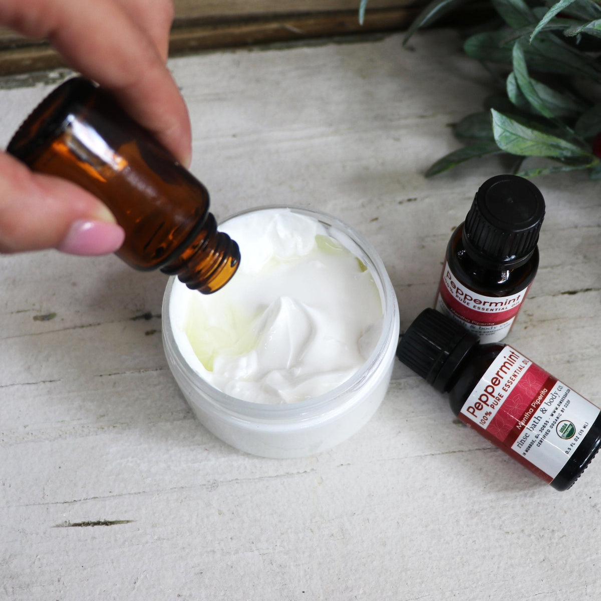 Essential oil dripping from an amber bottle into a jar of white cream, with Peppermint oil bottles nearby