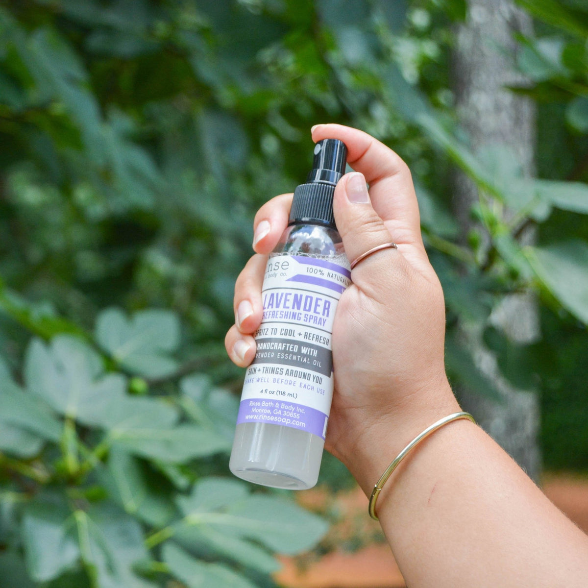 Hand holding a Lavender refreshing spray bottle in front of green fig leaves