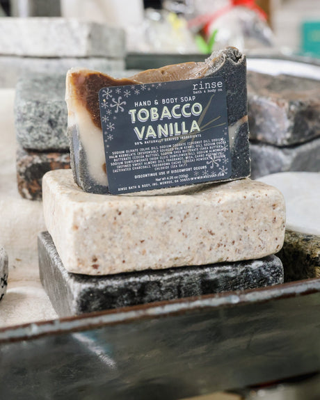 The Tobacco Vanilla hand and body bar resting on stone in a workshop — sweet tobacco, vanilla and cedar in a cocoa-butter base. Rinse Bath & Body Co.