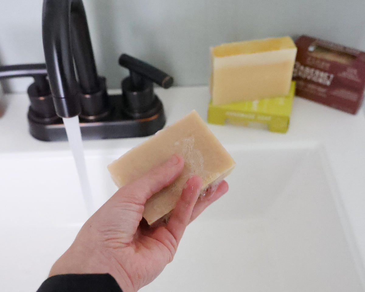 Hand holding Champagne soap in a running sink stream — lime and Pumpkin boxes on the counter