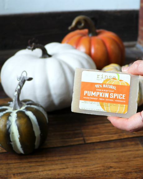 A hand holding the Pumpkin Spice bar in front of heirloom white and green pumpkins on weathered wood — nutmeg, clove and brown sugar, the bar that makes an ordinary shower smell like the first cold morning. Rinse Bath & Body Co.