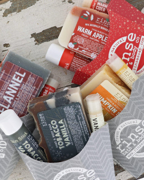 A curated bath/body gift assortment from Rinse & Body Co. arranged in a gray paper tray on a distressed white wood surface.