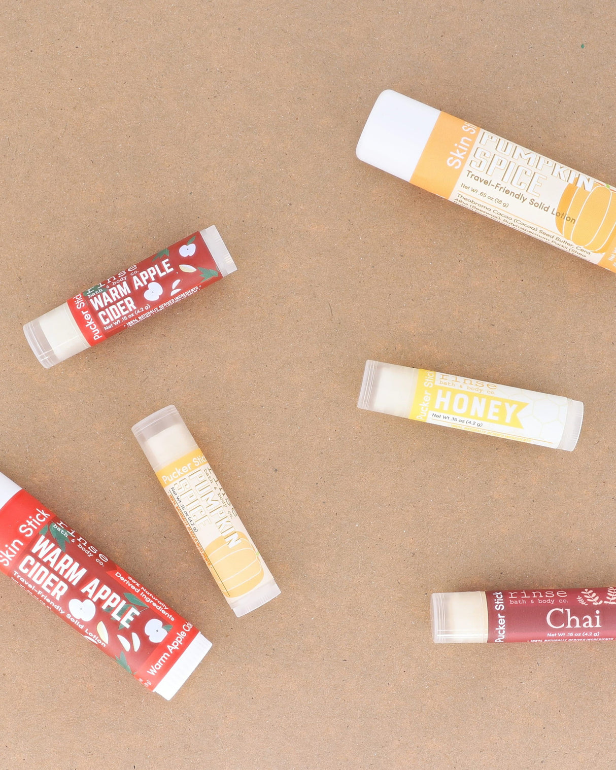 Warm Apple Cider lip balms scattered across warm kraft paper with the season's other flavours — spiced apple, clove and brown sugar for chapped autumn lips. Rinse Bath & Body Co.