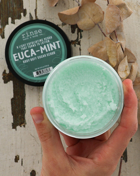 Hand holding a container of Euca-Mint body scrub with a visible brand label in the background.