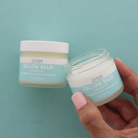Hand holding an open jar of unscented Tallow Balm with another jar in the background on a teal background