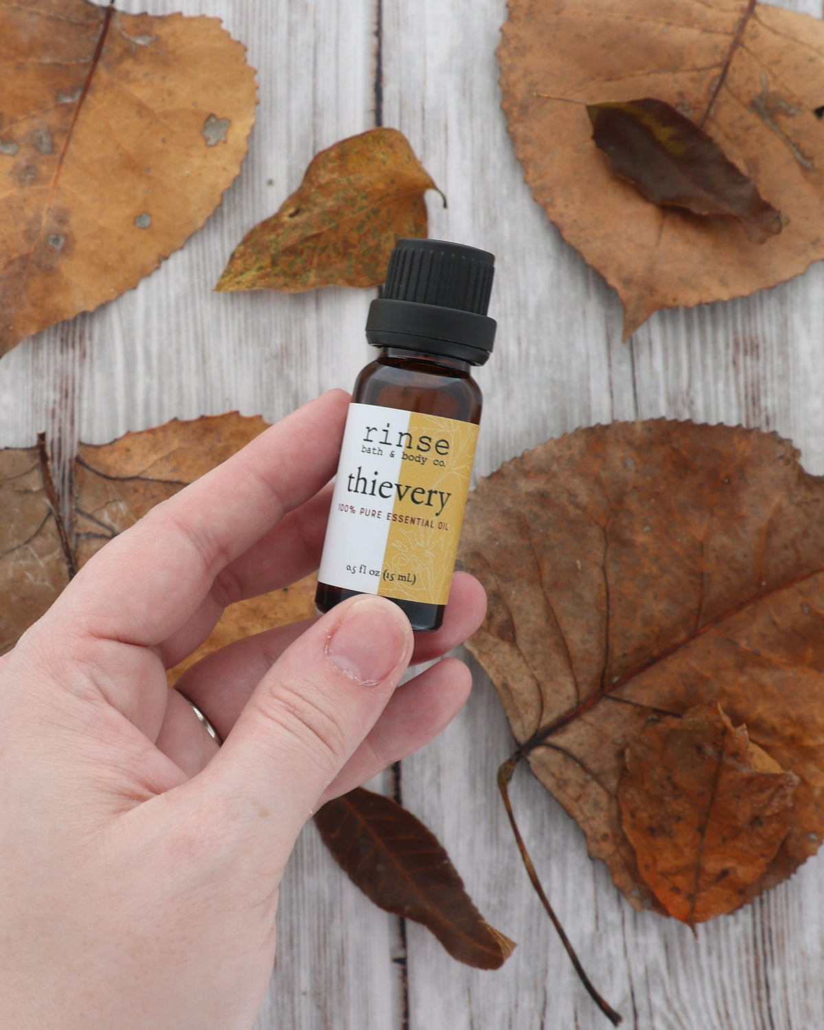 Hand holding a Thievery essential oil bottle with its gold label facing out above dried leaves on white wood