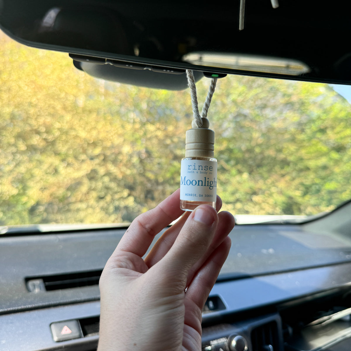 Hand holding the Moonlight car diffuser on its cord below a rearview mirror, autumn trees outside