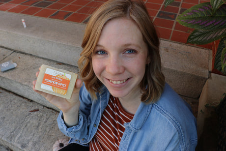 A woman outdoors smiling as she holds a bar of Pumpkin Spice soap — warm autumn spice, the gift that reads as fall the moment it's opened. Rinse Bath & Body Co.