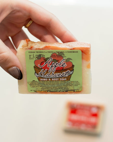 A hand holding the Apple Harvest bar with its orchard-print label facing out — crisp red apple and warm cinnamon, the soap that makes an ordinary shower feel like a fall morning. Rinse Bath & Body Co.