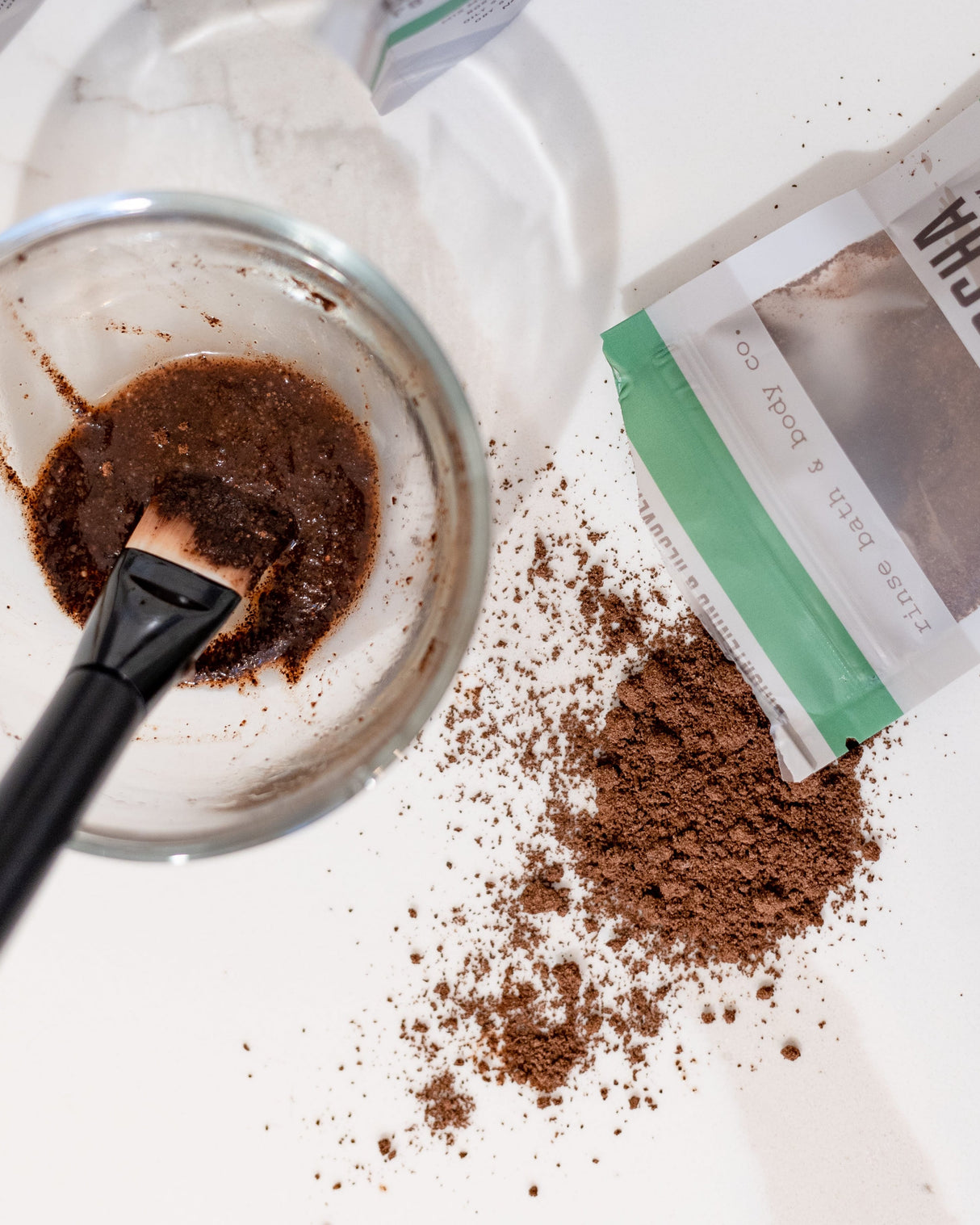 A mask brush resting in a bowl of mixed Cafe Mocha Face Mask, the pouch and loose coffee grounds beside it. Rinse Bath & Body Co.