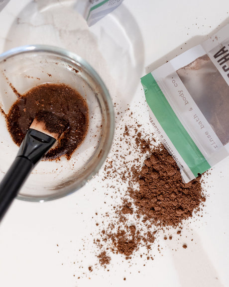 A mask brush resting in a bowl of mixed Cafe Mocha Face Mask, the pouch and loose coffee grounds beside it. Rinse Bath & Body Co.