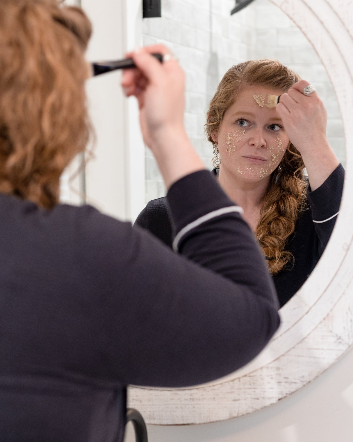 A woman smoothing Oatbrasion Face Mask onto her cheek at a round bathroom mirror. Rinse Bath & Body Co.