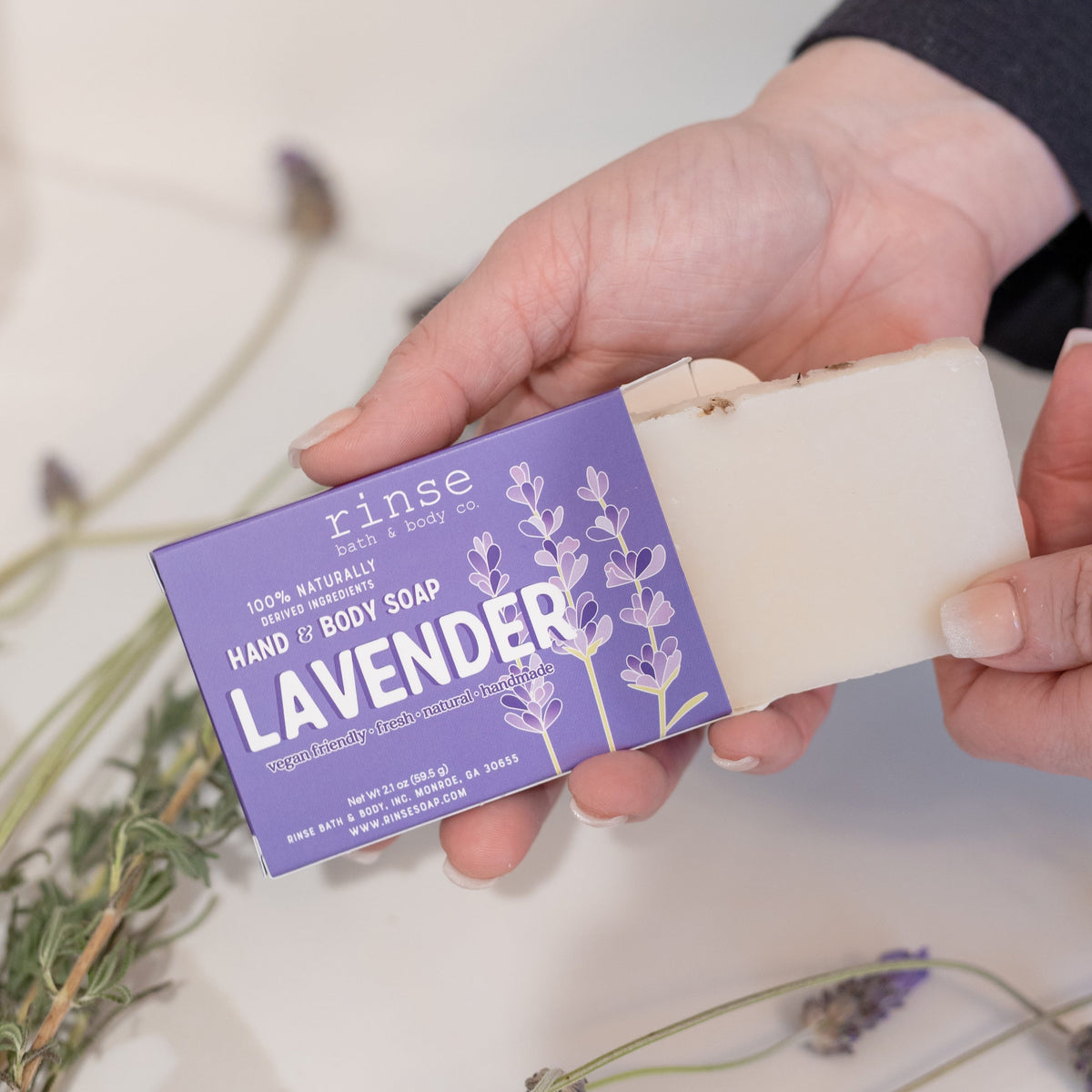Hands sliding creamy Lavender soap from its purple box beside dried lavender sprigs — bright white surface