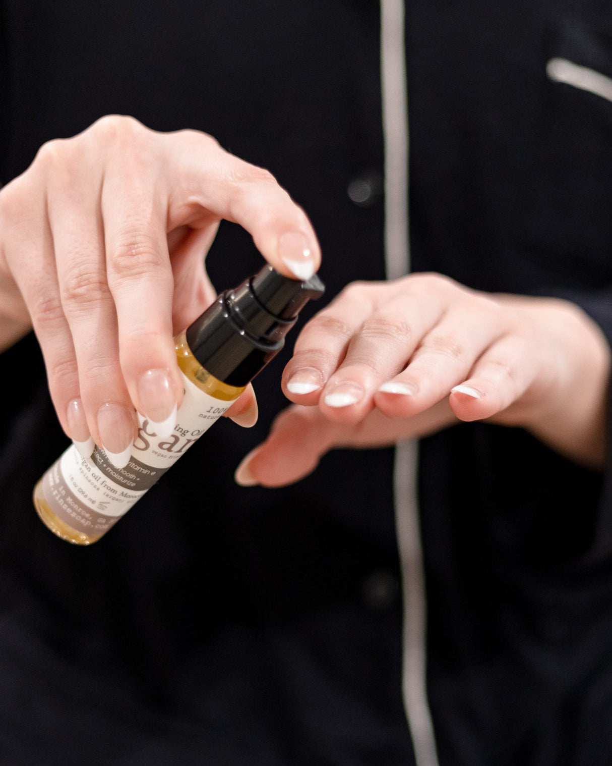 Argan oil dispensed from its glass dropper into an open palm against a dark shirt, a single amber drop suspended at the tip. Rinse Bath & Body Co.