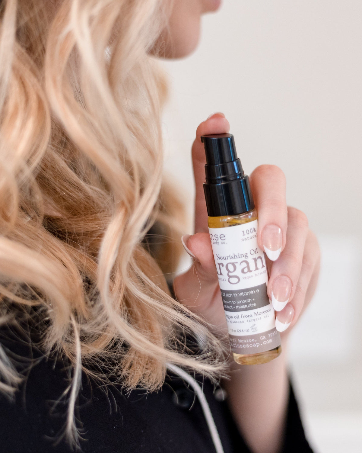 A woman with long blonde waves holding the Rinse Argan oil bottle beside her hair, thumb on the dropper — a multi-use oil for skin, nails and hair. Rinse Bath & Body Co.