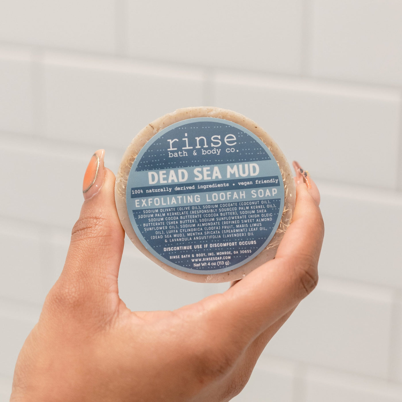 Two hands holding the round Dead Sea Mud Loofah bar with its blue label facing the camera — mineral mud and a loofah face for exfoliating and detoxing in one bar. Rinse Bath & Body Co.