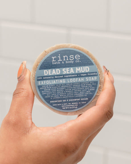 Two hands holding the round Dead Sea Mud Loofah bar with its blue label facing the camera. Rinse Bath & Body Co.