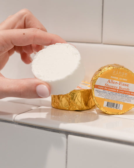 A hand holding a white Aller-Ease shower steamer puck beside its gold foil wrapper on white subway tile