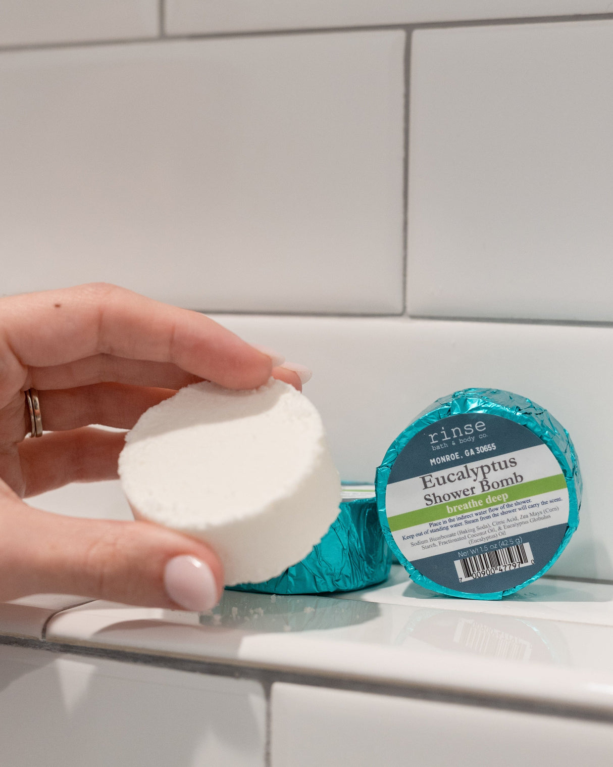 A hand holding a white Eucalyptus shower steamer puck beside its teal foil wrapper on white tile
