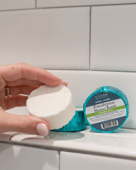 A hand holding a white Eucalyptus shower steamer puck beside its teal foil wrapper on white tile