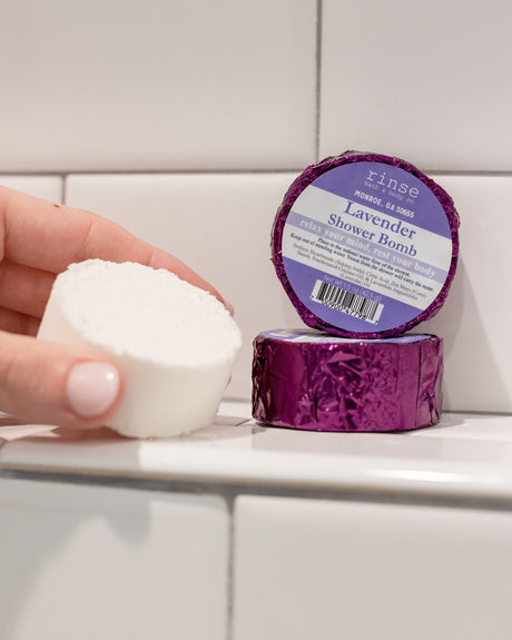 A hand holding a white Lavender shower steamer puck beside its purple foil wrapper on white subway tile
