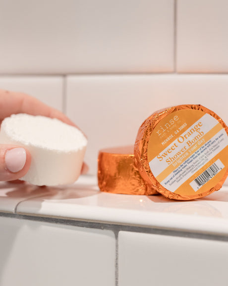 A hand holding a white Sweet Orange shower steamer puck beside its orange foil wrapper on white tile