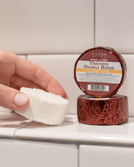 A hand holding a white Thievery shower steamer puck beside its maroon foil wrapper on white subway tile