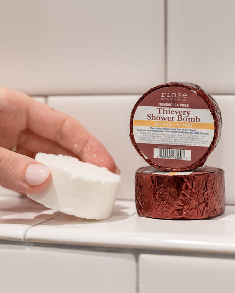A hand holding a white Thievery shower steamer puck beside its maroon foil wrapper on white subway tile — warm cinnamon, clove and eucalyptus steam for cold season. Rinse Bath & Body Co.