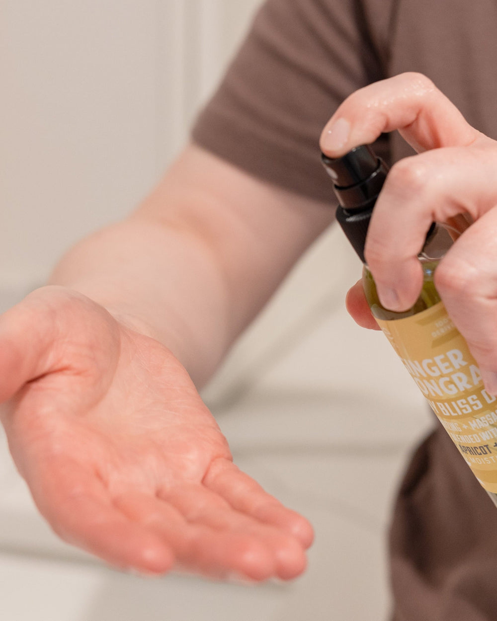 A hand sprays Ginger Lemongrass Body Bliss Oil into an open palm indoors — bright lemongrass over warm ginger in a squalane and apricot oil that absorbs fast. Rinse Bath & Body Co.
