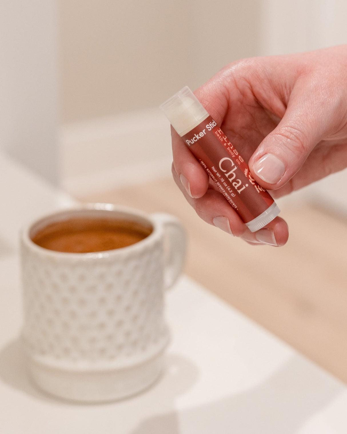 A hand holding the Chai Tea Latte lip balm beside a mug of spiced milk tea — cinnamon, cardamom and vanilla for soft winter lips. Rinse Bath & Body Co.