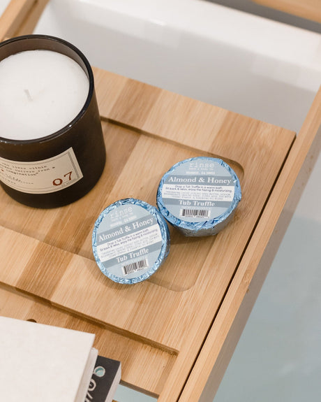 Two Almond Honey Tub Truffles on a bamboo bath caddy beside a lit candle and a folded cloth, bath below. Rinse Bath & Body Co.