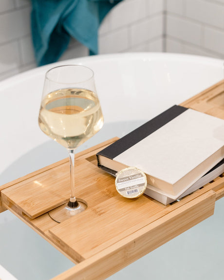 A Sweet Vanilla Tub Truffle on a bamboo bath caddy with a glass of white wine, a book and a candle, beside a filled freestanding tub. Rinse Bath & Body Co.