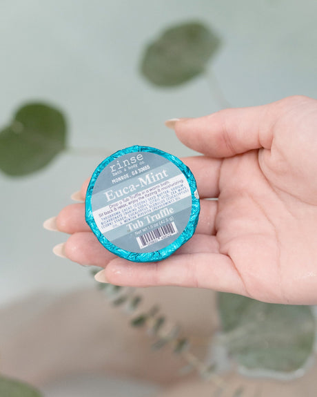 An open palm holding a teal foil-wrapped Euca-Mint Tub Truffle above a filled bath, eucalyptus stems trailing behind. Rinse Bath & Body Co.