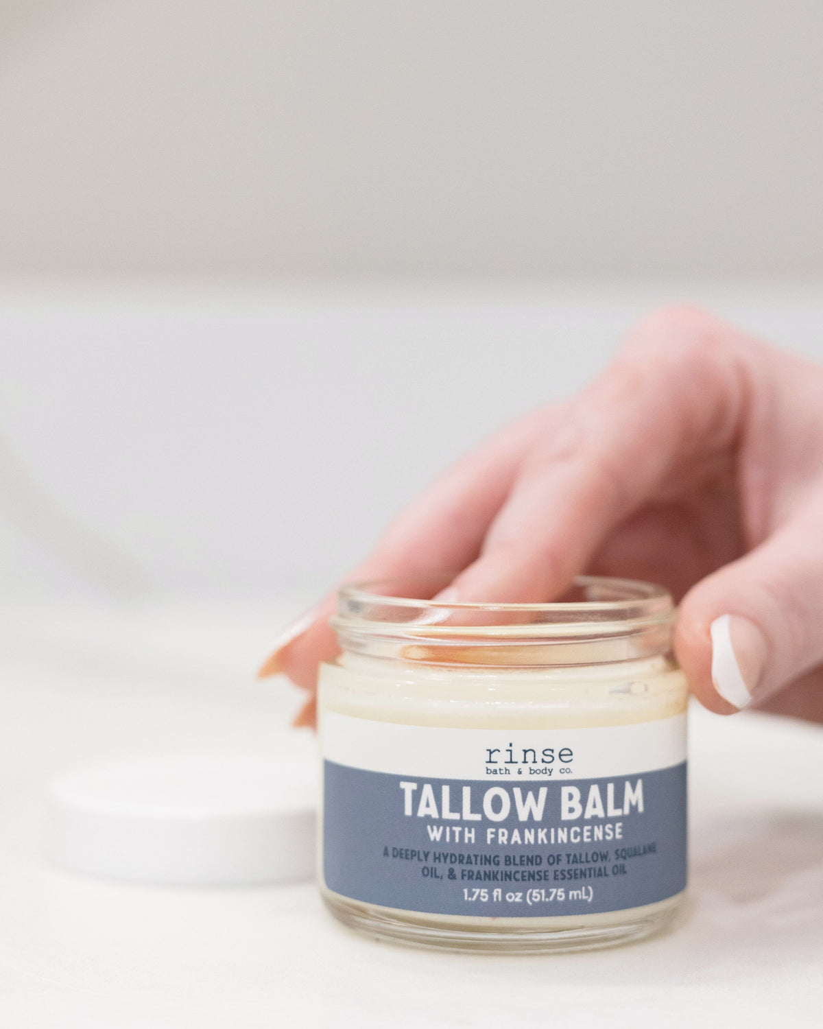 Two hands lifting the lid from a glass jar of Frankincense Tallow Balm, blue label facing out — grass-fed beef tallow and frankincense for deep repair on dry patches. Rinse Bath & Body Co.