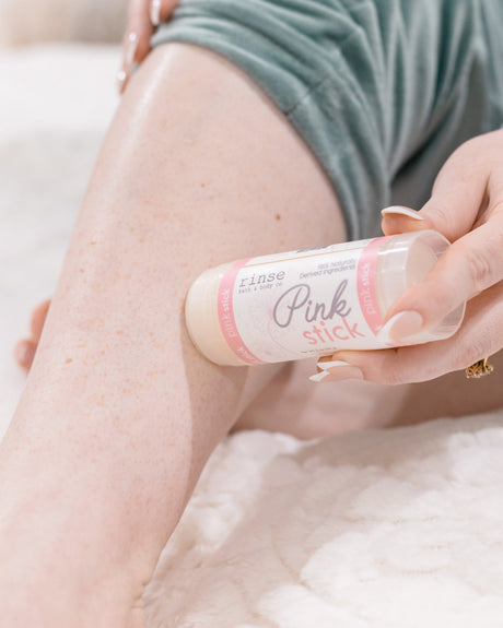 A hand gliding the Pink Skin Stick along a bare leg — waterless butter-rich moisture applied straight to skin. Rinse Bath & Body Co.