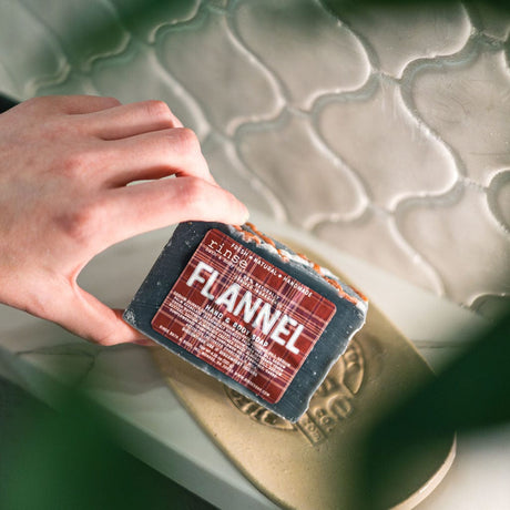 A hand lifting the Flannel bar from a stoneware dish against sage-green tile — cedar and sweet tobacco, the bar that turns a weekday shower into a cabin morning. Rinse Bath & Body Co.