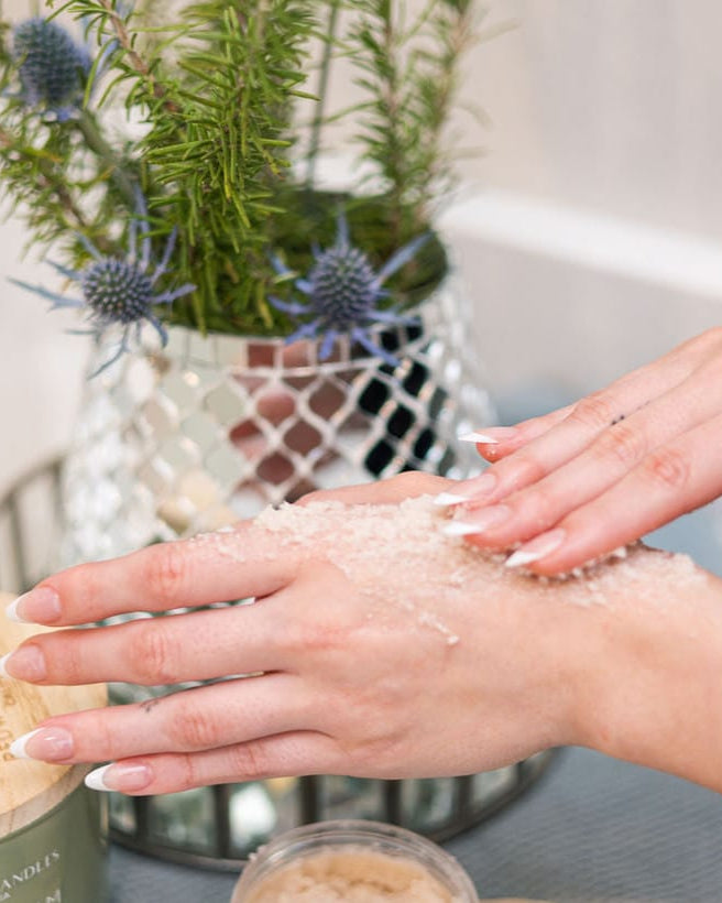 Hands working granular sugar scrub over the back of a hand beside a silver mesh planter of rosemary and blue thistle, an open jar at the edge of frame — the scrub step, before it rinses to a lotion finish. Rinse Bath & Body Co.