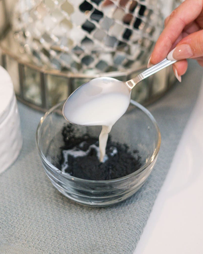 A spoonful of mask paste poured into a glass bowl of black charcoal powder, mixing CharClay Face Mask by hand. Rinse Bath & Body Co.
