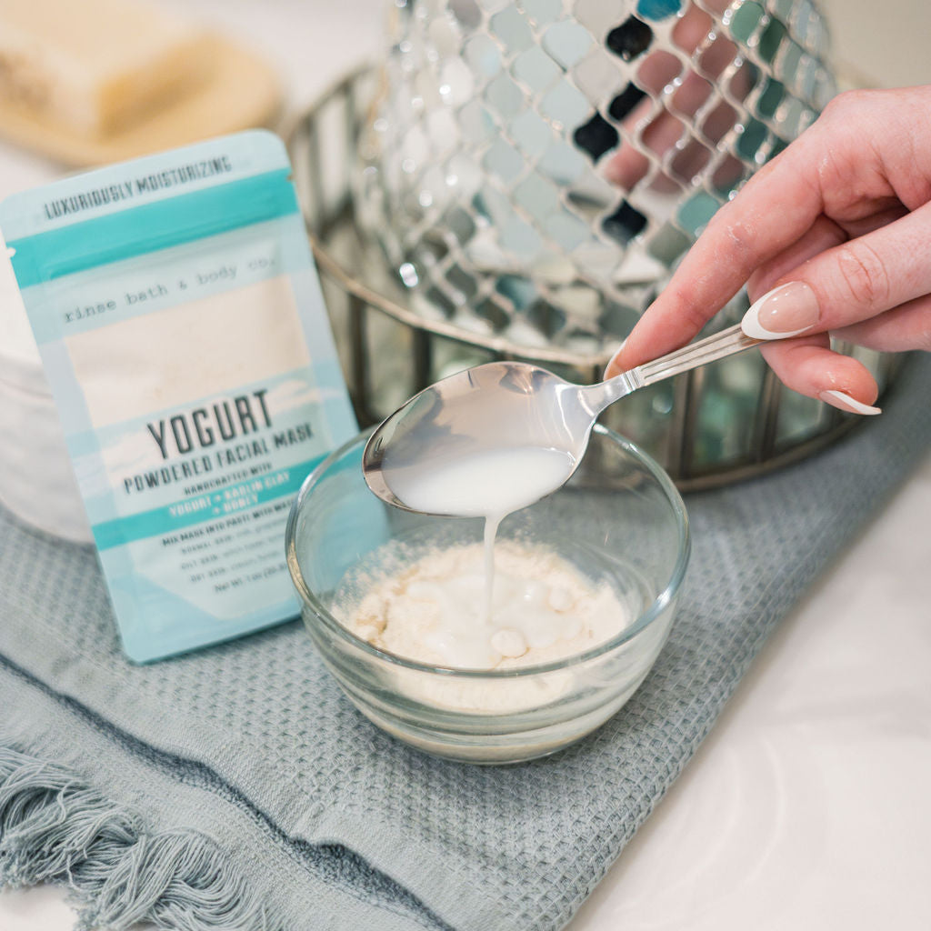A spoonful of Yogurt Face Mask powder lifted from a bowl, the pouch resting on grey linen beside it. Rinse Bath & Body Co.