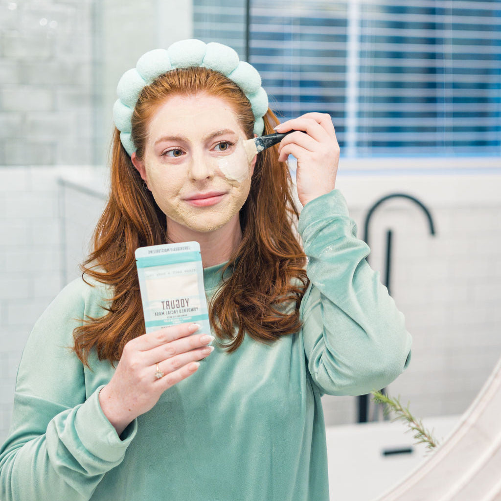 A woman in a headband applying Yogurt Face Mask at the bathroom mirror. Rinse Bath & Body Co.