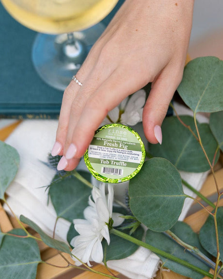 A hand setting a green-labelled Fresh Fig Tub Truffle among eucalyptus and white blooms on a wooden bath tray — green, a little sweet, not remotely floral. Rinse Bath & Body Co.