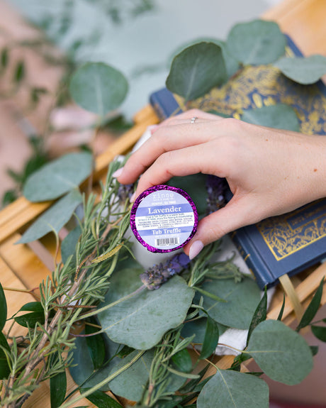 A hand placing a purple foil-wrapped Lavender Tub Truffle among fresh lavender and eucalyptus on a wooden bath tray — three lavender baths, boxed. Rinse Bath & Body Co.