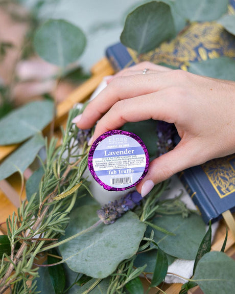 A hand reaching in to place a purple foil-wrapped Lavender Tub Truffle among fresh lavender and eucalyptus on a wooden bath tray, cloth-bound books beside it. Rinse Bath & Body Co.