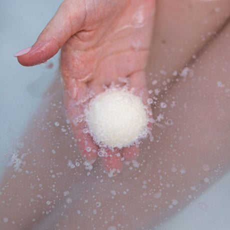 A hand holding a white cocoa butter Tub Truffle just under the surface of a warm bath, fizzing as it dissolves and releasing the cocoa butter into the water. Rinse Bath & Body Co.