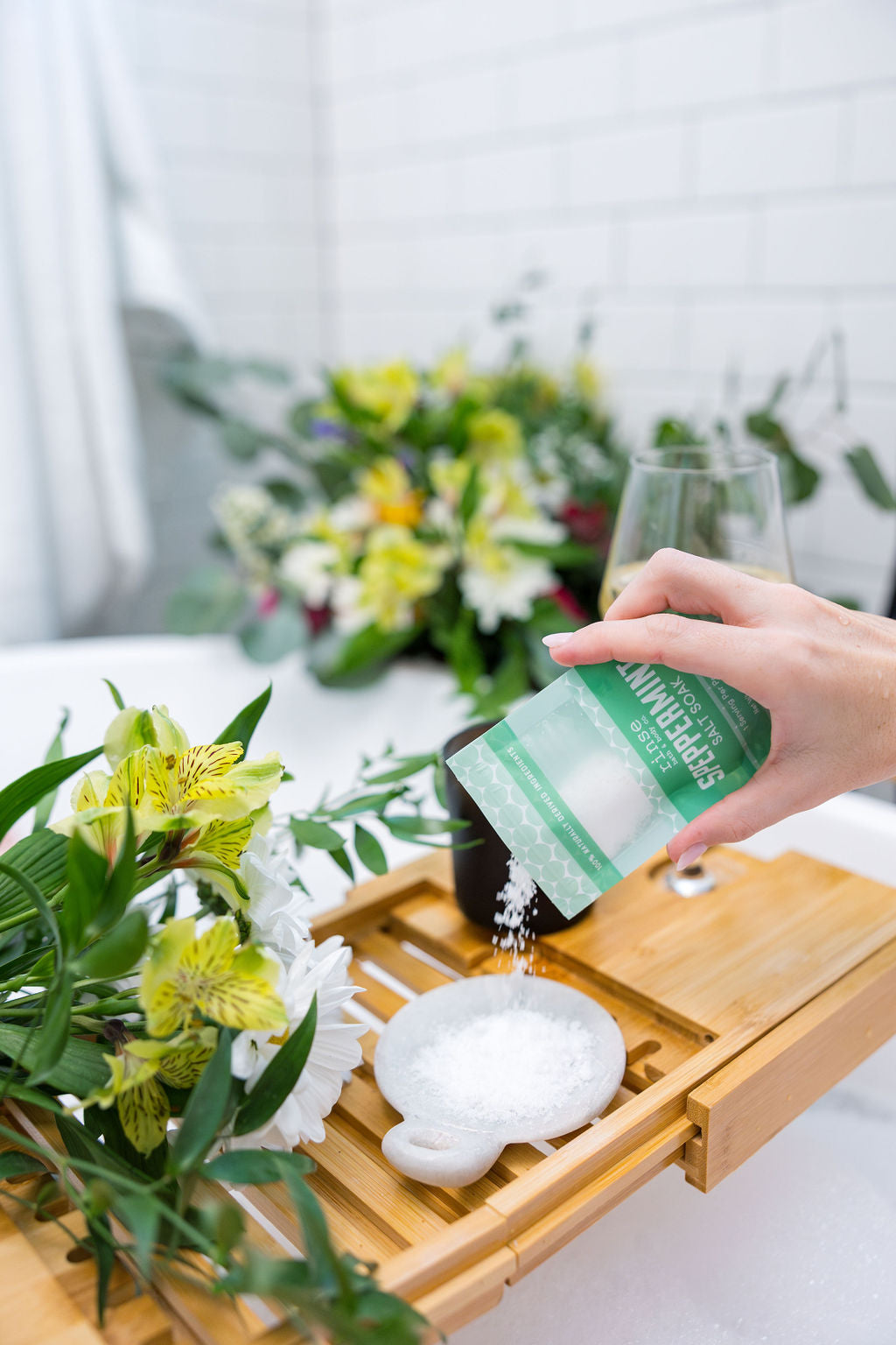 Hand tipping Speppermint Salt Soak into a white dish on a bamboo caddy — yellow flowers candle and glass beside