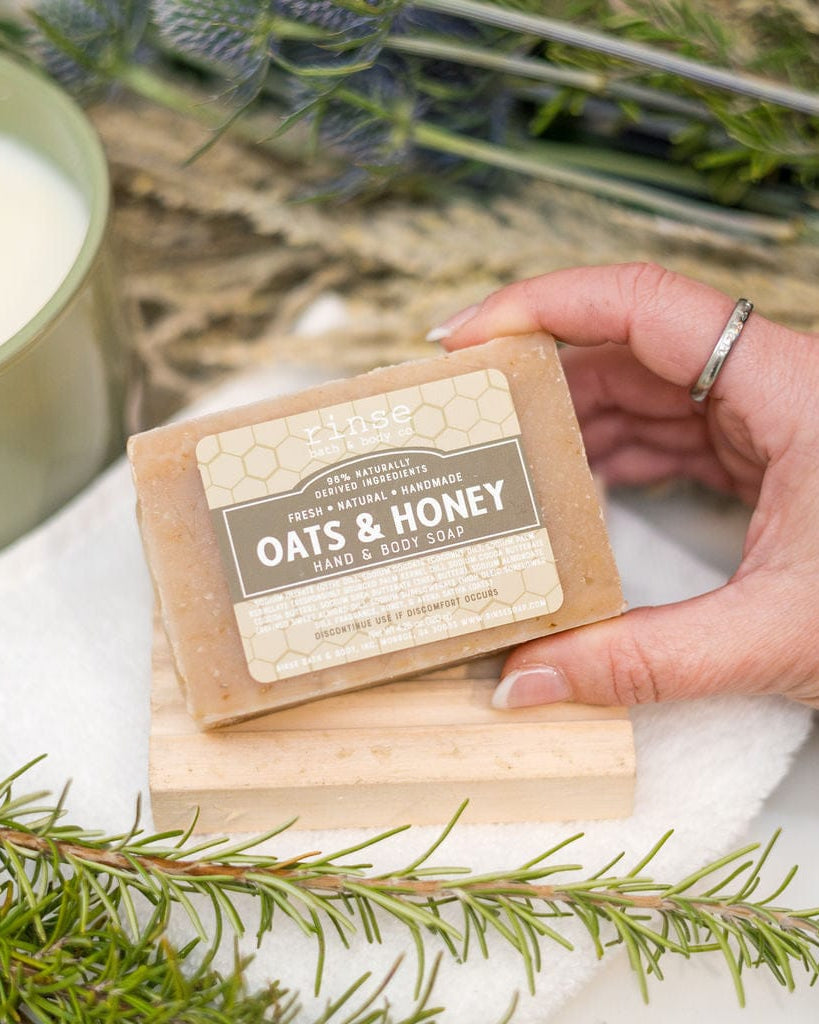 A hand holding the Oats & Honey bar beside fresh rosemary and a lit candle — colloidal oatmeal and raw honey, the bar people reach for when skin needs calming. Rinse Bath & Body Co.