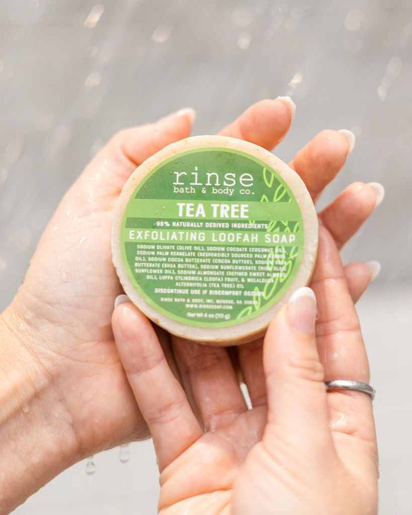 Two cupped hands hold the round Tea Tree Exfoliating Loofah Soap with its green label facing out, water beading on tile behind — clarifying tea tree in a scrubbing loofah bar. Rinse Bath & Body Co.