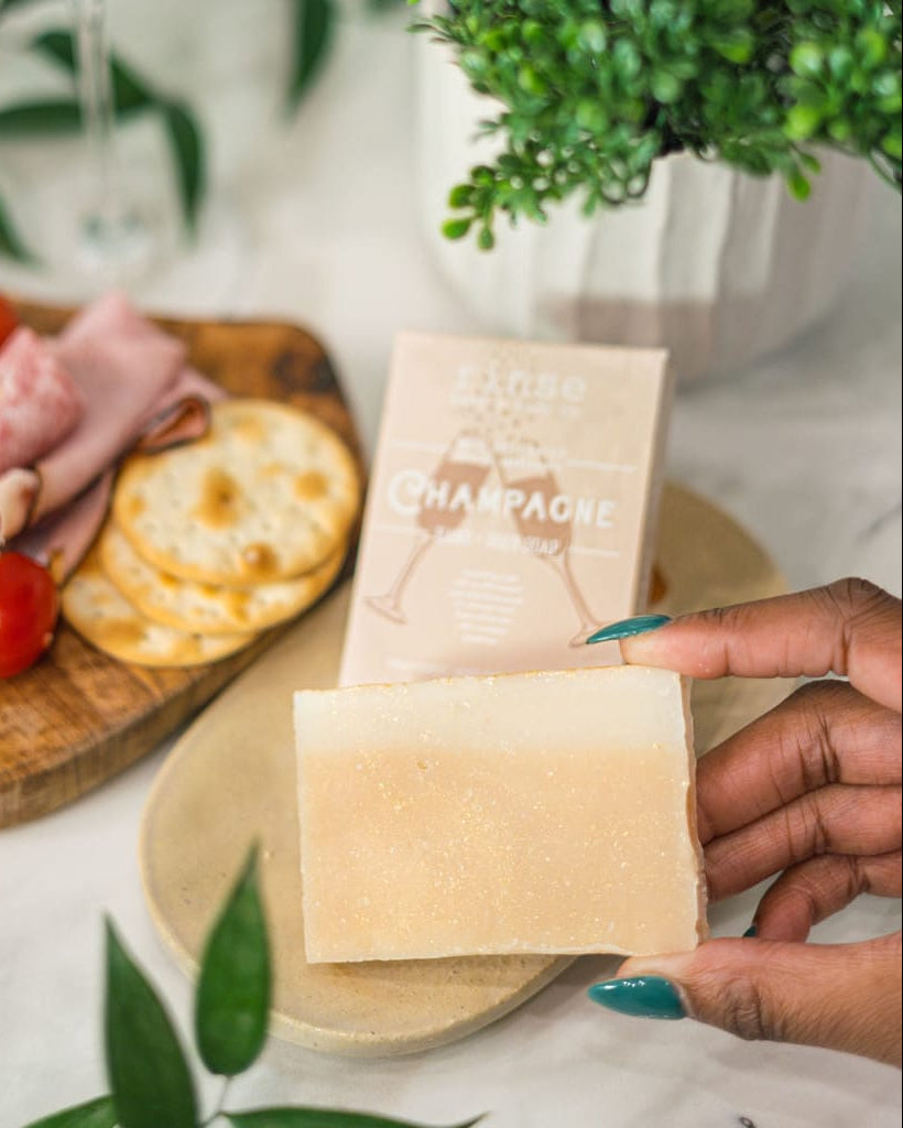 Hand holding Champagne soap beside charcuterie and greenery in bright daylight — bubbly sparkling notes for a festive rinse