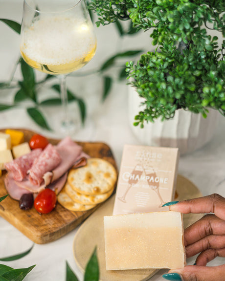 Teal-nailed hand holding Champagne soap beside a bubbly flute — charcuterie board and plant in bright light
