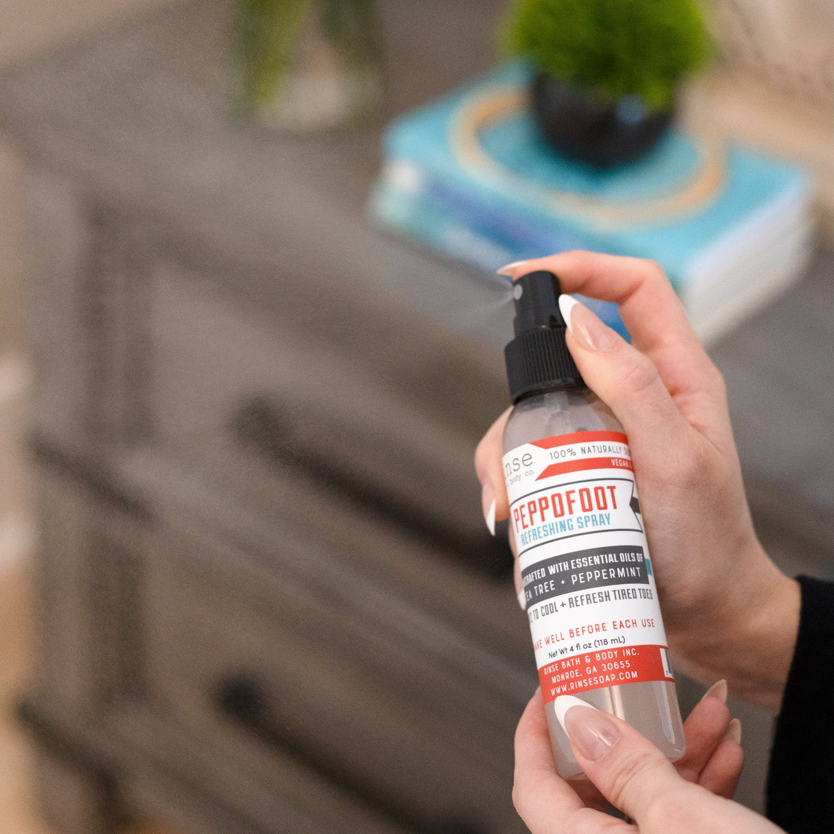 A hand holding Peppofoot Refreshing Spray beside a bedside table with a plant and stacked books — the last step before bed for tired feet. Rinse Bath & Body Co.